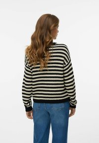 Vrouw met lang golvend haar, gekleed in een zwart-wit gestreepte trui en blauwe spijkerbroek, gezien van achteren tegen een egale achtergrond.