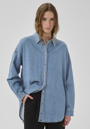 Naiste helepruuni juustega, kellel on seljas lahtine sinisest denimist nööbitav särk ja mustad püksid, üks käsi taskus, neutraalse taustaga.