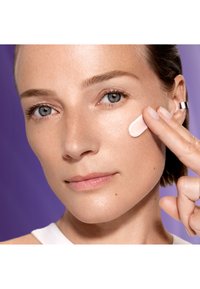 Un gros plan d'une personne appliquant une crème rose clair sur sa joue, sur un fond violet avec une texture de peau lisse et un maquillage naturel.