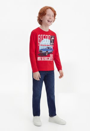 Rødhåret dreng smilende, iført en rød langærmet Shelby GT500 bilgrafiktrøje, blå jeans og hvide sneakers, stående mod en hvid baggrund.