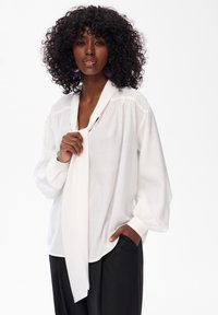 Biała, lekkie bluzka z wiązaniem przy szyi, długimi bufkami i marszczonymi akcentami. Połączona z czarnymi spodniami.