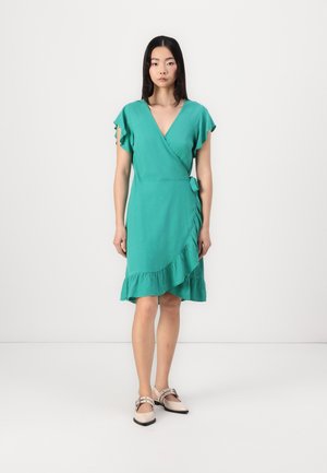 Abito a portafoglio teal con maniche a volant e orlo a strati. Presenta una cintura laterale per la regolazione. La modella indossa ballerine beige.
