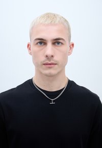 Čierne tričko s okrúhlym výstrihom, strieborný náhrdelník s malým horizontálnym príveskom, krátke blond vlasy, modré oči, neutrálny pozadie.