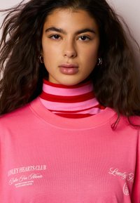 Roza majica s belim besedilom, oblečena preko rdečega in rožnatega prugastega puloverja z visokim ovratnikom. Ima okrogel izrez in kratke rokave.