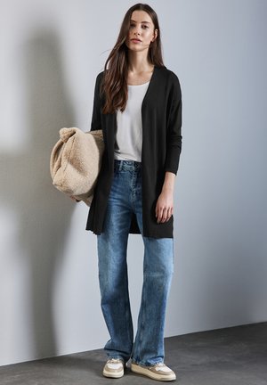 Zwarte open voorkardigan over een witte T-shirt, gecombineerd met lichte blauwe flared jeans en beige sneakers. Een zachte beige tas wordt vastgehouden.