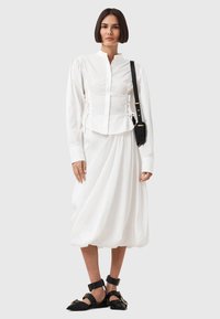 Blusa bianca con maniche lunghe e colletto alto, abbinata a una gonna bianca fluida. Sandali neri con fibbie completano il look.