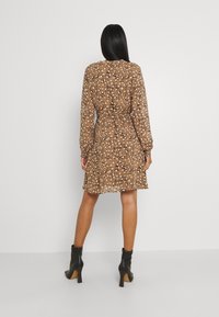 Mujer con cabello negro largo, vestida con un vestido marrón de estampado hasta la rodilla y botas de tacón negro hasta el tobillo, de pie contra un fondo blanco liso.