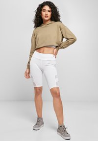 Īss olīvu krāsas kapuču džemperis kopā ar baltām riteņbraukšanas šortiem ar tīkliņa paneļiem. Modelis valkā pelēkas augstās sporta kurpes. Vienkāršs, mūsdienīgs sporta apģērbs.