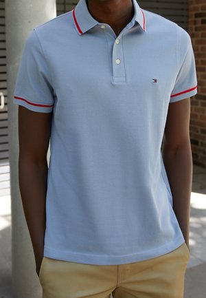 Person trägt hellblaues Poloshirt mit roten Streifen am Kragen und an den Ärmeln, beige Hose, steht mit den Händen in den Taschen im Freien.