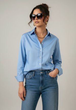 Vrouw met oversized zwarte zonnebril, lichtblauw overhemd met knopen, gouden ketting en oorbellen, en blauwe spijkerbroek met één hand in de zak.