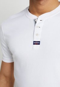 Bílá bavlněná košile s krátkým rukávem Henley se třemi knoflíky, malým logem Superdry na hrudi.