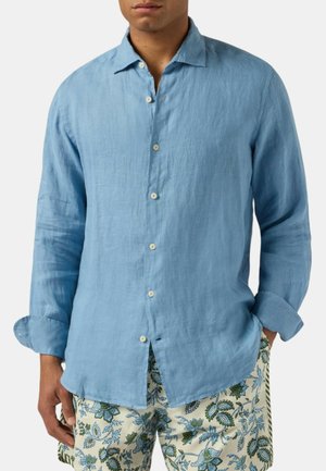 Uomo che indossa una camicia di lino azzurro chiaro a maniche lunghe con bottoni abbinata a pantaloncini bianchi con fantasia floreale con foglie verdi e blu.