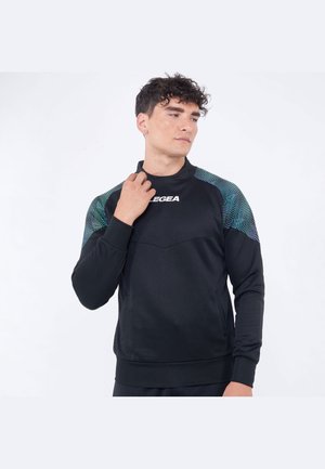 Giovane uomo con maglia sportiva nera a maniche lunghe con spalle decorate, guarda di lato tenendo il colletto con una mano.
