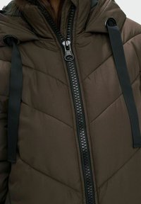 Bruine gewatteerde jas met capuchon, voorzien van een zwarte rits en een gestructureerd chevronpatroon. Zwarte trektouwen accentueren het ontwerp.