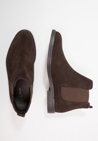 Botas de ante marrón hasta el tobillo con paneles laterales elásticos, punta redondeada y una tira en la parte trasera. Suela de goma plana con detalle de costura.