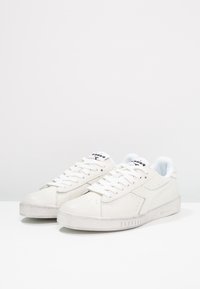 Vita lädersneakers med texturerad yta, platta snören, perforerade detaljer och gummisula. Har logotyp på tungan och minimalistisk design.