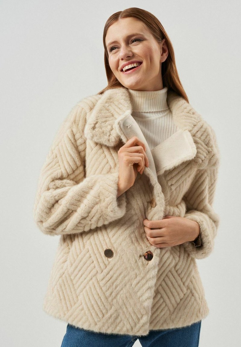 Beżowa kurtka z sztucznego futra w wzór diamentów, z dużym kołnierzem i dwoma guzikami z przodu. Posiada teksturowany materiał i luźny krój.