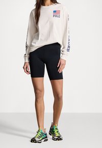 Långärmad beige bomullsskorta med amerikanska flaggan och "POLO"-logotyp; matchad med svarta cykelbyxor och färgglada träningsskor.