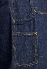 Lähedalt vaade tumedast sinisest teksakangast, millel on kahekordse õmblusega oranž nõel ja ruudukujuline tasku, mille kohal on väike silmus.