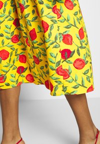Falda amarilla con un vibrante patrón de frutas rojas y hojas verdes. El dobladillo es redondeado y la tela parece ligera con una textura suave.
