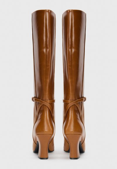 Botas de cuero marrón hasta la rodilla con una textura suave, que presentan un diseño de tiras cruzadas y tacones gruesos. Estética limpia y minimalista.