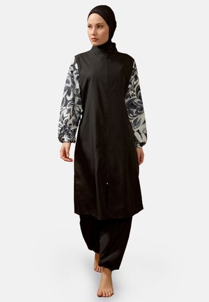 Woman barefoot wearing a long black zip-up dress with black and white floral patterned sleeves and a black headscarf walking forward.