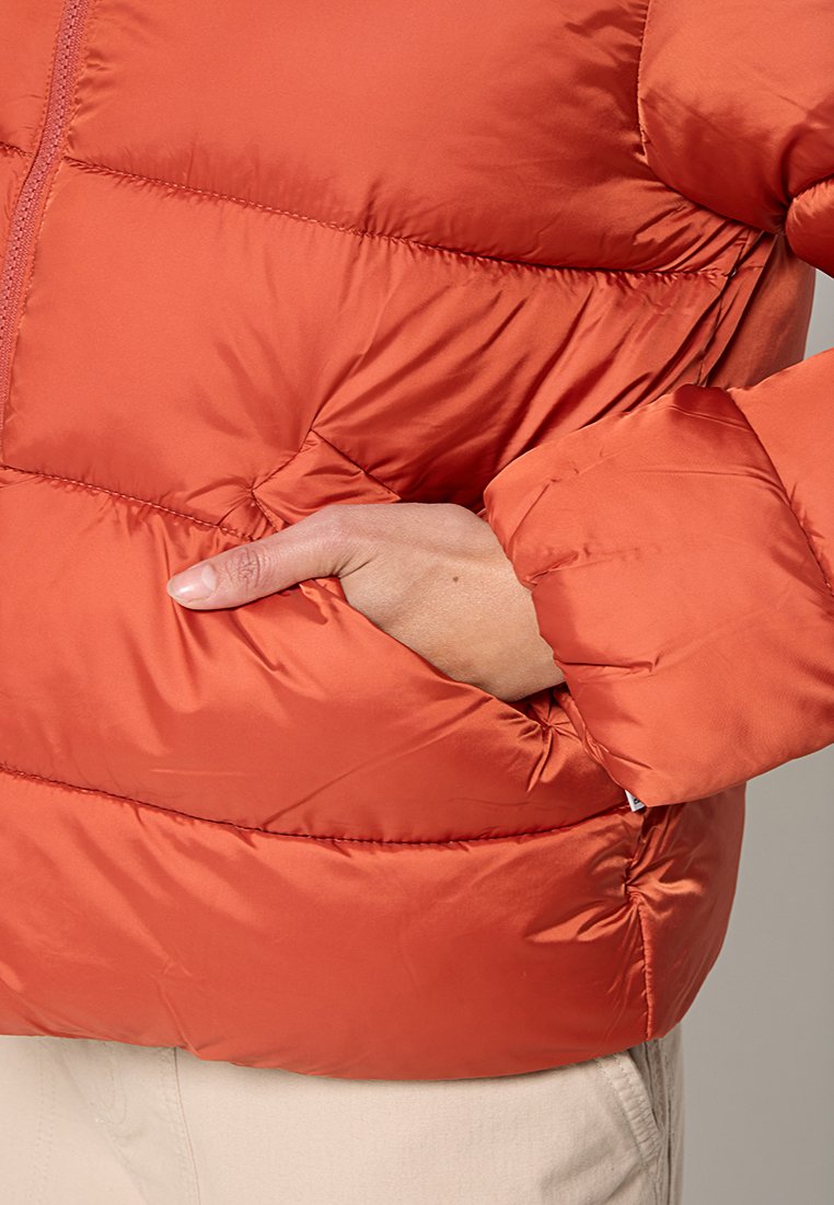 Die Hand einer Person steckt in der Tasche einer orangefarbenen Daunenjacke mit Reißverschluss, während sie hellfarbige Hosen trägt.