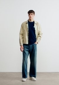 Νεαρός άνδρας στέκεται μπροστά σε απλό λευκό τοίχο φορώντας μπεζ μπουφάν, μπλε πουκάμισο, φαρδιά μπλε τζιν και λευκά αθλητικά παπούτσια, με το ένα χέρι στην τσέπη.