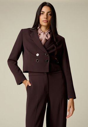 Vrouw met lang donker haar draagt een donker cropped dubbele knoopsluiting blazer over bijpassende high-waisted broek en een lichtroze strikblouse.