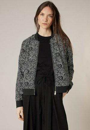 Femme aux longs cheveux bruns portant une veste zippée à motif noir et blanc sur un haut noir et un pantalon noir à taille nouée.