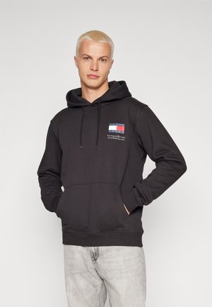 Sudadera negra con un bolsillo canguro frontal, capucha con cordón y un pequeño parche de logo de color en el pecho. Textura de tela suave.