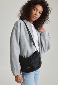 Sac en bandoulière noir matelassé avec quincaillerie dorée et une sangle de marque, présenté sur un fond gris clair avec une personne portant une veste grise.