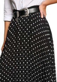 Jupe plissée noire à pois blancs, avec une large ceinture noire ornée d'une boucle en argent ciselé et des détails en tissu texturé.