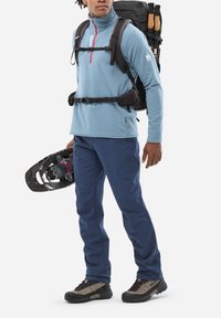 Pull bleu clair en polaire avec une fermeture éclair rouge, pantalon bleu marine, bottes de randonnée noires et beiges, portant une raquette noire et un sac à dos noir.