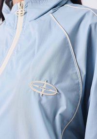 Giacca a vento azzurro chiaro con piping bianco, colletto alto e un patch rotondo sul petto. Tessuto liscio e leggero.