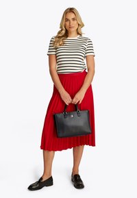 Black leather handbag with rectangular shape, two handles, and silver hardware. Model wears a striped shirt and a pleated red skirt.