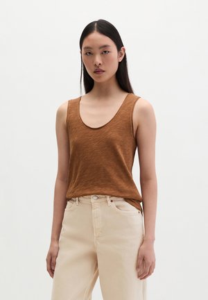 Jeune femme aux longs cheveux foncés portant un haut marron sans manches et un pantalon beige taille haute, debout devant un fond uni.