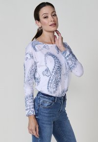 Mujer posando con la mano cerca del cuello, vistiendo una camisa blanca de manga larga con estampado paisley azul y jeans de mezclilla azules, contra un fondo liso.