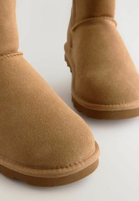 Világosbarna színű, domború orrú, varrott varrású és gumitalpú bokacsizma, amely puha textúrát és egyszerű dizájnt mutat.