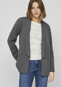 Blazer gris léger avec une fermeture ouverte, coupe ajustée et deux poches ; porté par-dessus un pull blanc texturé et un jean bleu.