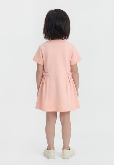 Tüdruk seisab seljaga vaataja poole, seljas roosad lühikeste varrukatega kleit, mille külgedel on voltimised, ja valged libisevad jalanõud, taust on ilma mustrita.