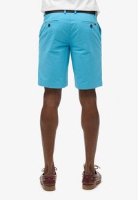 Pantalones cortos azul brillante, material de algodón, corte clásico, dos bolsillos traseros con botones negros, llevados con zapatos de barco de cuero marrón.