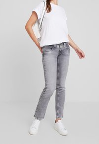 Jeans de mezclilla gris con un ligero desgastado, diseño de cinco bolsillos y acento verde en la cinturilla. Combinados con una camiseta blanca y zapatillas blancas.