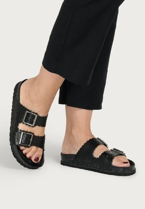 Feet wearing black sandals with two buckled straps and floral cut-out patterns, paired with cropped black pants against a plain background.