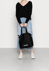 Persoon in een zwarte trui en versleten lichtblauwe jeans, met een zwarte Levi's tote bag, staand tegen een plain witte muur.