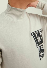 Person som bär en beige rippad polotröja med stora marinblå och vita "WS" bokstäver på bröstet, hand som rör vid nacken.