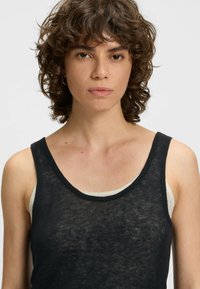 Schwarzes ärmelloses Tanktop mit Rundhalsausschnitt, aus leichtem, durchscheinendem Stoff, über einem weißen Tanktop getragen. Keine bemerkenswerten Muster oder Metallteile.