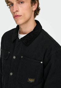 Črna bombažna jakna z ovratnikom iz korduroja, sprednjim zapiranjem na gumbe, dvema žepoma na prsih, srebrno kovinsko opremo in označbo blagovne znamke na spodnjem žepu.