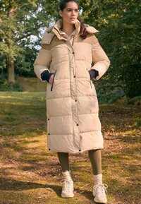 Lang beige dunjakke med høj krave, sorte accenter og trykknapper. Bæres over olivengrønne leggings og hvide sneakers i en skovagtig omgivelser.