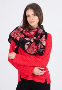 Floral gebreide sjaal in zwart met rode en beige bloemmotieven. Zachte textuur, om de schouders gedrapeerd, gecombineerd met een gestructureerde rode trui.